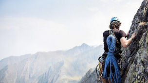 Eine Person klettert an einer Felswand und trägt dabei einen Helm und Kletterausrüstung, einschließlich eines Seils, das um ihren Körper gewickelt ist. Im Hintergrund sind Berge unter einem klaren Himmel sichtbar.