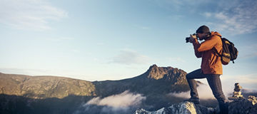 Eine Person fotografiert eine Berglandschaft, während sie einen orangefarbenen Anorak und einen Rucksack trägt. Der Himmel ist klar und die Sonne scheint.