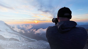 Eine Person fotografiert einen Sonnenaufgang über einer Berglandschaft, während Wolken den Himmel bedecken.