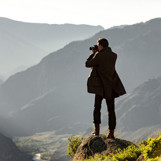 Eine Person steht auf einem Felsvorsprung und fotografiert ein weitläufiges Gebirgstal. Verschachtelte, nebelige Bergketten erstrecken sich in die Ferne; weiches Gegenlicht und lange Schatten betonen die Tiefe der Landschaft.