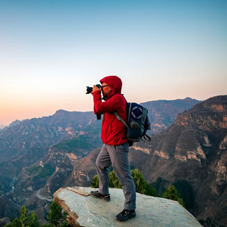 Eine Person mit roter Jacke und Rucksack steht auf einer Felskante und fotografiert die weite, zerklüftete Berglandschaft. Die Gipfel und Täler erstrecken sich bis zum Horizont, der Himmel leuchtet in sanften Pastellfarben von Sonnenauf- oder -untergang.
