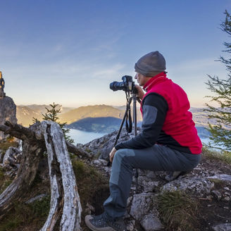 Ein Landschaftsfotograf kniet auf einem felsigen alpinen Hang neben einem Stativ, trägt rote Weste und graue Mütze. Vor ihm liegen zerklüftete Felsen, ein umgestürzter Baumstamm und Nadelbäume; darunter schlängelt sich ein See durch sonnenbeschienene Bergtäler unter klarem blauem Himmel.