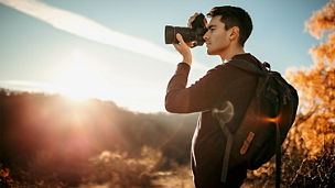 Eine Person mit Rucksack fotografiert im Gegenlicht der tief stehenden Sonne, seitlich im Profil, das Gesicht ist unscharf/abgedeckt. Warmes Herbstlicht, goldene Laubfärbung und Lens Flares prägen die Szene; im Hintergrund liegen Sträucher und ein leicht hügeliges Gelände. Die Stimmung wirkt wie zur goldenen Stunde.
