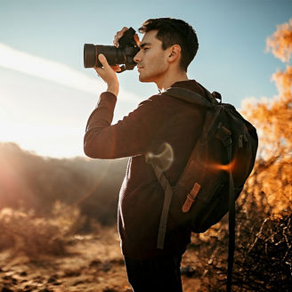 Eine Person mit Rucksack fotografiert im Gegenlicht der tief stehenden Sonne, seitlich im Profil, das Gesicht ist unscharf/abgedeckt. Warmes Herbstlicht, goldene Laubfärbung und Lens Flares prägen die Szene; im Hintergrund liegen Sträucher und ein leicht hügeliges Gelände. Die Stimmung wirkt wie zur goldenen Stunde.