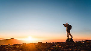 Eine einzelne Wanderin bzw. ein Wanderer steht auf einem felsigen Grat bei Sonnenaufgang und fotografiert den Horizont. Der Himmel ist klar, warmes orangefarbenes Licht liegt über der Landschaft, die Person mit Rucksack ist als Silhouette vor der aufgehenden Sonne zu sehen.