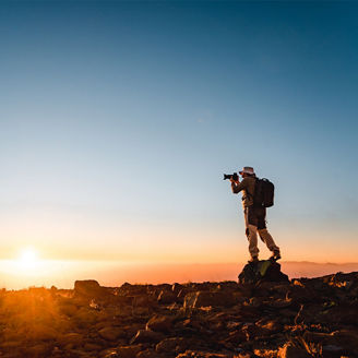 Eine einzelne Wanderin bzw. ein Wanderer steht auf einem felsigen Grat bei Sonnenaufgang und fotografiert den Horizont. Der Himmel ist klar, warmes orangefarbenes Licht liegt über der Landschaft, die Person mit Rucksack ist als Silhouette vor der aufgehenden Sonne zu sehen.