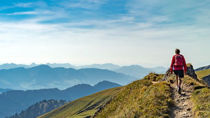 Das Bild zeigt eine Person auf einem schmalen Wanderweg entlang eines grünen Bergrückens mit Bergketten im Hintergrund. Der Himmel ist klar und blau.
