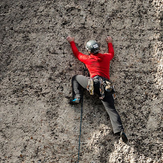 Das Bild zeigt einen Kletterer in roter Kleidung und Helm, der eine steile Felswand erklimmt. Er ist mit Kletterausrüstung gesichert und konzentriert sich darauf, den nächsten Griff zu erreichen.