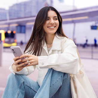 Waiting for the bus and for love. Bright cheerful young woman of Turkish mixed race in white casual clothes with a charming smile holding a phone in the street at public transportation stop