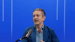 Person speaking into a microphone in a studio, seated against a blue background with vertical light accents