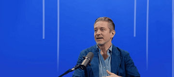 Person speaking into a microphone in a studio, seated against a blue background with vertical light accents