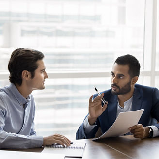 Two diverse serious young male business partners discussing contract terms, agreement text, marketing financial report, reviewing paper document at workplace table together