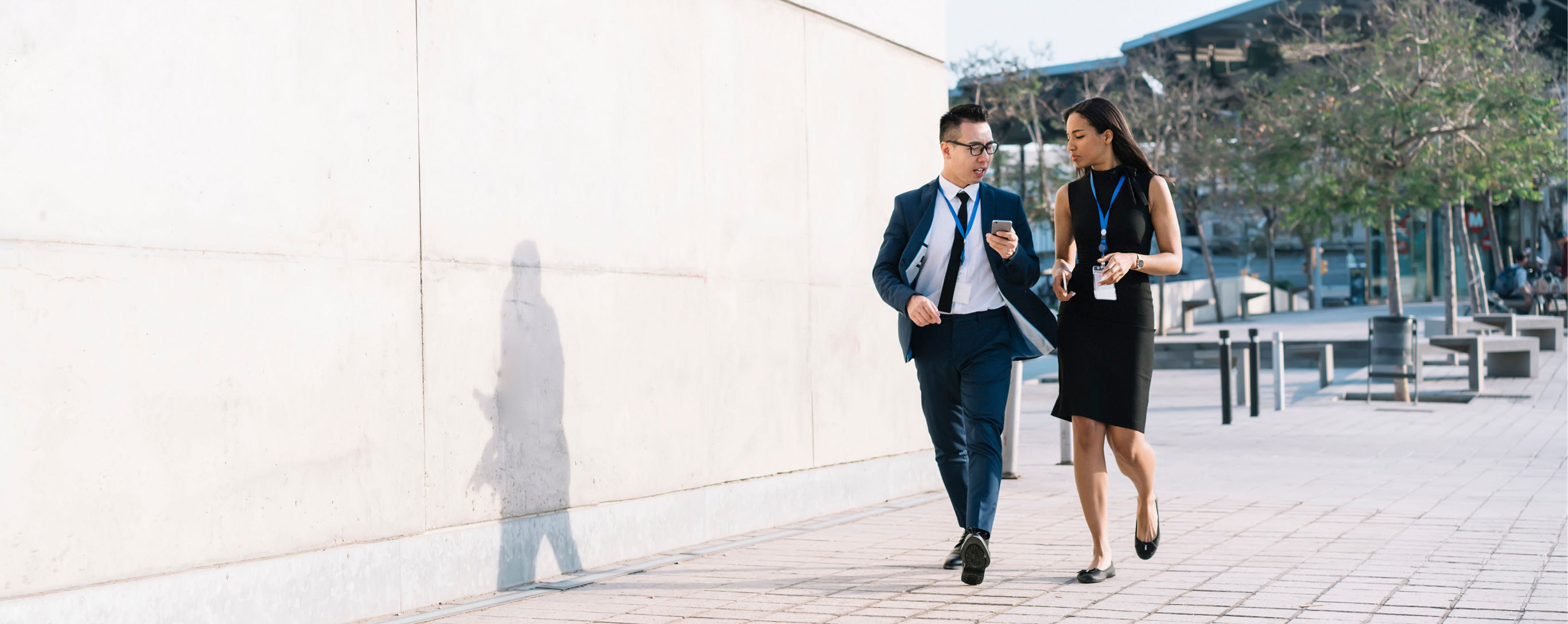 Young elegant diverse man and woman in formal clothes and id badges having coffee and sharing phone while walking on street