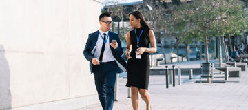 Young elegant diverse man and woman in formal clothes and id badges having coffee and sharing phone while walking on street