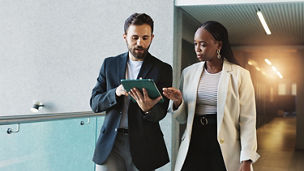 Two professionals reviewing information on a tablet in an office hallway.