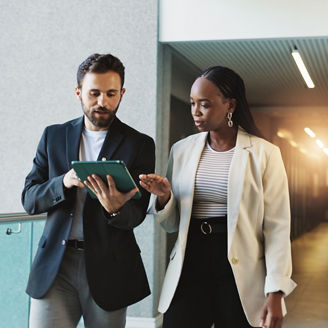 Two professionals reviewing information on a tablet in an office hallway.