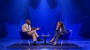 Two speakers sit in chairs on a stage with a Canadian maple leaf in the background