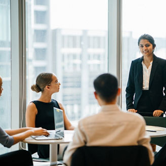 An Asian Indian businesswoman is having a conversation with her colleagues in a conference room and looking forward for their project. She's wearing a black blazer and white blouse with a black skirt.