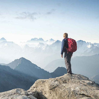 Das Bild zeigt eine Person mit einem roten Rucksack, die auf einem Felsen steht und in die Ferne blickt. Im Hintergrund erstreckt sich eine beeindruckende Berglandschaft mit mehreren Gipfeln, die von Nebel und Dunst umgeben sind. Der Himmel ist leicht bewölkt, und die Szene vermittelt ein Gefühl von Freiheit und Abenteuer in der Natur.