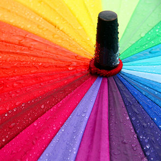 Das Bild zeigt die Nahaufnahme eines bunten Regenschirms, der in einem Regenbogenmuster gestaltet ist. Die Farben verlaufen von Rot über Orange, Gelb, Grün, Blau, bis hin zu Violett. Auf der Oberfläche des Schirms sind zahlreiche Wassertropfen sichtbar, die auf den Stoff gefallen sind. Der zentrale Stab des Schirms ist ebenfalls zu sehen.