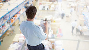 Person reviewing inventory on a clipboard in a warehouse with shelves and stored goods.