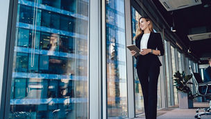 Caucasian female entrepreneur dressed in formal wear going near panoramic window in office, confident business woman with modern digital tablet looking away during working day in enterprise