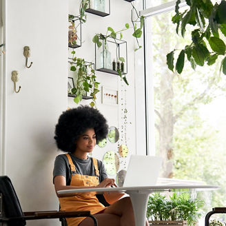 Happy stylish hipster African American female student with Afro hair using laptop computer sitting at table learning at home, in cozy modern cafe, remote studying, watching video, working in internet.