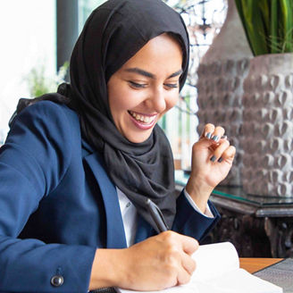 Happy businesswoman taking notes in coffee shop. Young Muslim business woman in hijab and office suit writing in notebook and laughing. Muslim business professional concept