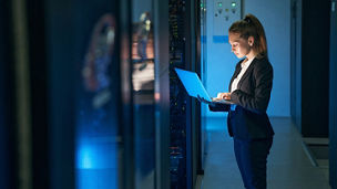 Woman IT specialist in elegant suit working on notebook computer in data center next to server racks