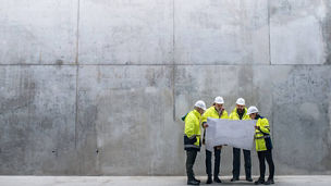 Group of workers in high-visibility clothing reviewing plans against a plain concrete wall